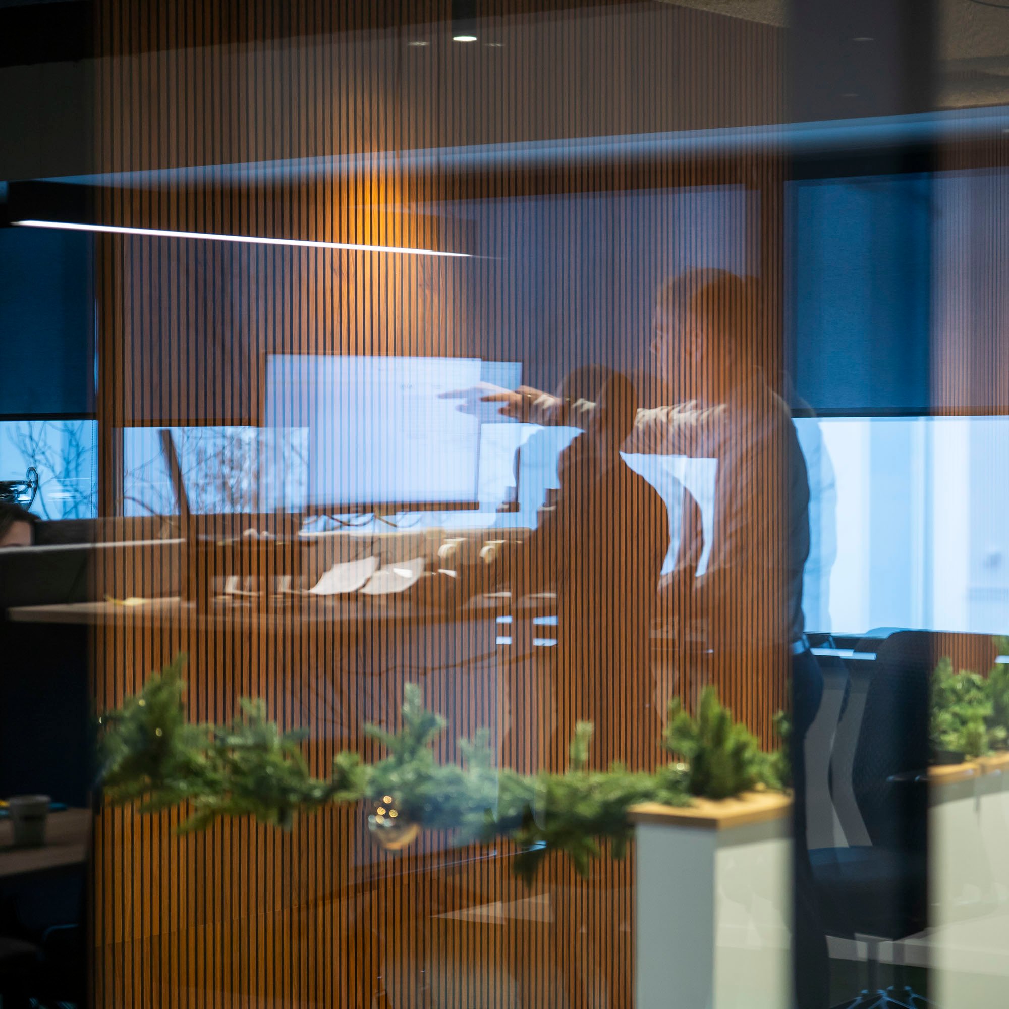 A blurry reflection of a person points at a computer monitor inside an office with wooden slat walls and foreground green foliage.