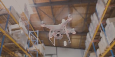 A white drone hovers in a warehouse aisle with tall shelves of stored goods.