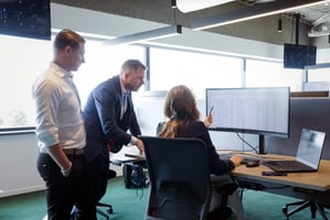 Three colleagues discuss data displayed on a curved monitor and laptop at an office desk. A woman points at the screen while two men observe.