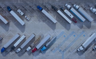 Semi-trucks and cars are parked in an aerial view of a large outdoor lot. Blue and white parking lines are on the ground, with blue numbers '12', '13', '23' faintly visible.
