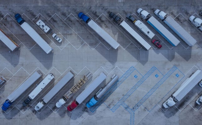 Semi-trucks and cars are parked in an aerial view of a large outdoor lot. Blue and white parking lines are on the ground, with blue numbers '12', '13', '23' faintly visible.