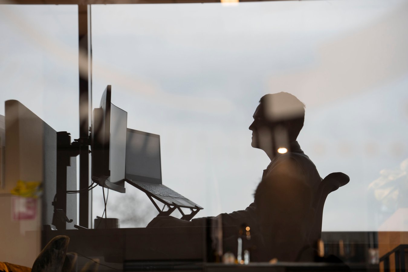 A person's silhouette sits at a desk with two monitors and a laptop on a stand, viewed through a reflective window against a bright sky.