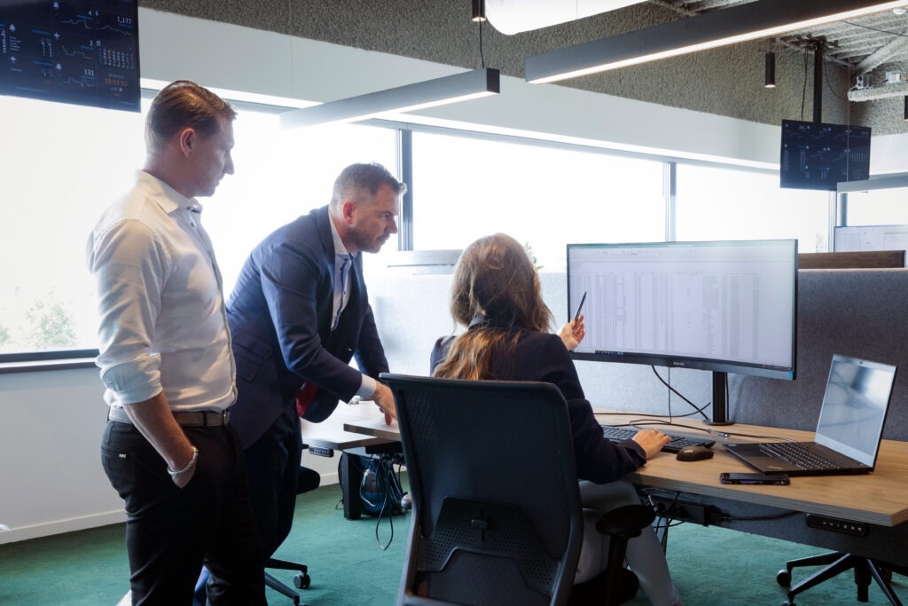 Three colleagues discuss data displayed on a curved monitor and laptop at an office desk. A woman points at the screen while two men observe.