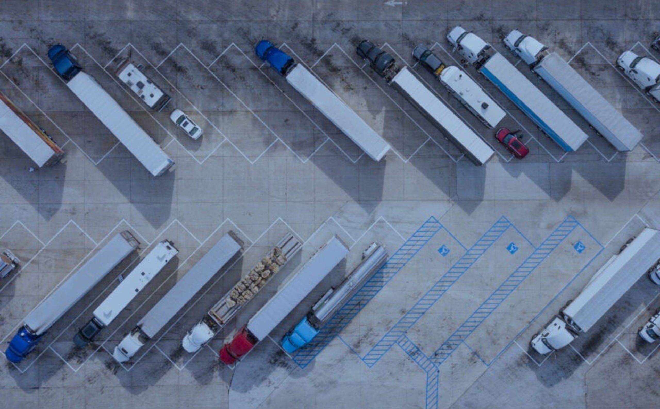 Semi-trucks and cars are parked in an aerial view of a large outdoor lot. Blue and white parking lines are on the ground, with blue numbers '12', '13', '23' faintly visible.